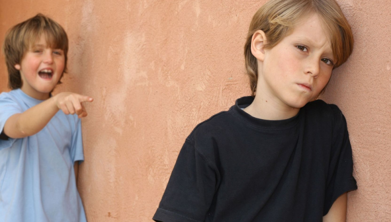 trauriger Junge lehnt an einer Mauer. Hinter ihm steht ein anderer JUnge, der ihn auslacht und mit dem Finger auf ihn zeigt.